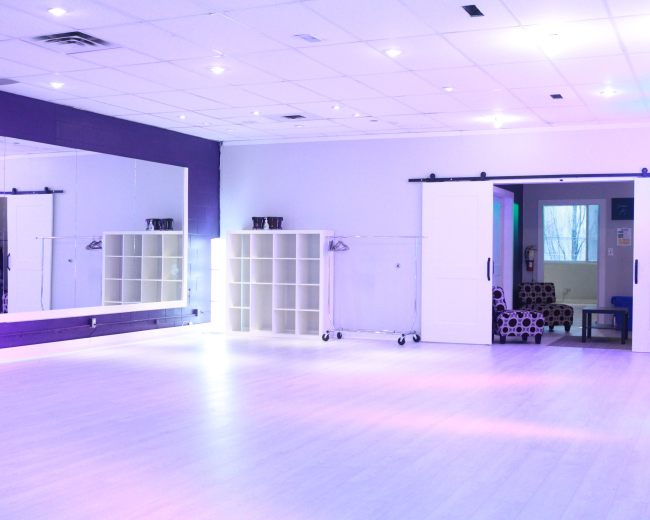 A brightly lit dance studio features a large mirrored wall, shelving unit, and a cozy seating area in the background.
