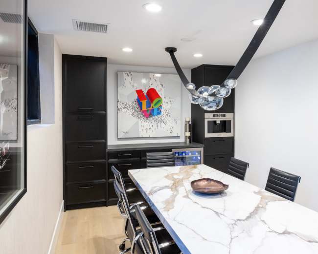 A modern dining area features a long marble table surrounded by black chairs, with a colorful art piece and a stylish light fixture overhead.