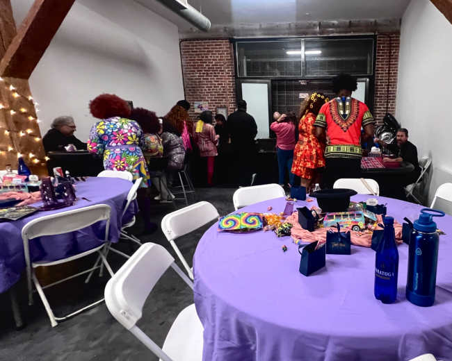 The image shows a gathering in a room with purple tablecloths on round tables, where individuals dressed in colorful attire are engaged in conversation.