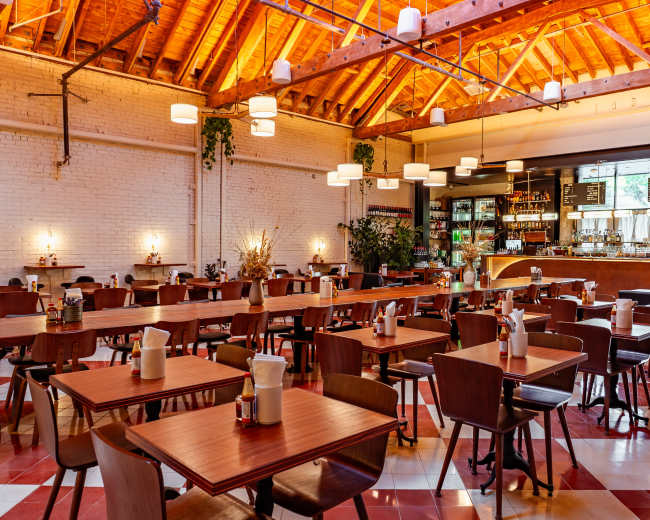 The image shows a spacious, modern restaurant with wooden tables and chairs arranged neatly, featuring a bar area with a variety of beverages in the background.