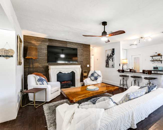 The living room features a white sofa, two armchairs, a wooden coffee table, and a mounted flat-screen TV above a decorative fireplace, next to a small dining area with barstools.