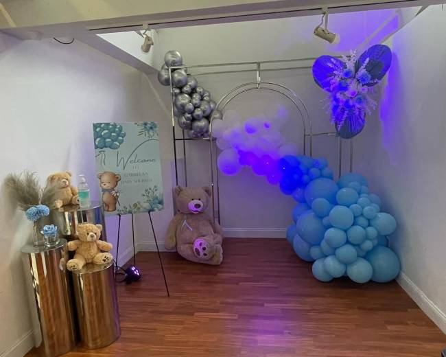 A decorated corner of a room featuring a balloon arch in shades of blue, stuffed bears, and a welcome sign.