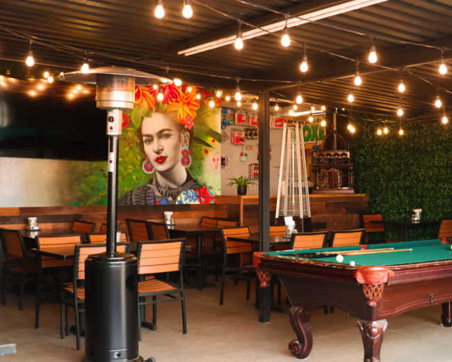 The scene features a vibrant outdoor dining area with a pool table, string lights, and a mural of a woman against a green backdrop.