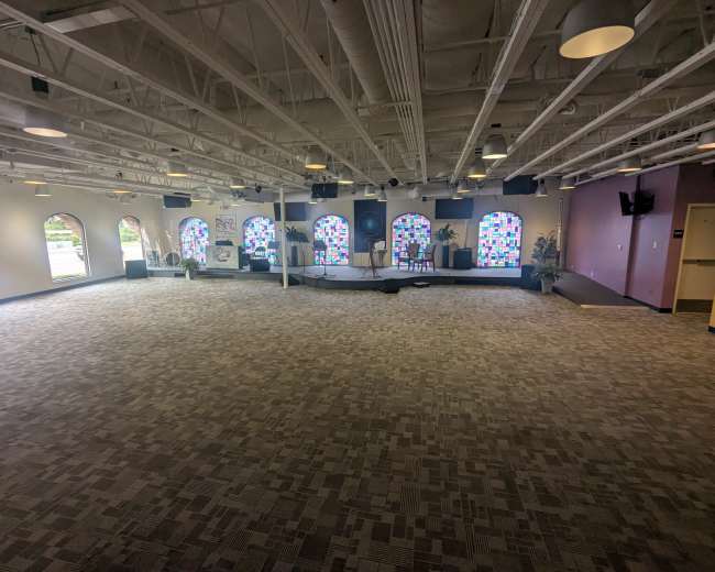 The image shows a spacious, empty room with patterned carpeting, large windows featuring colorful panes, and a stage area at the far end.