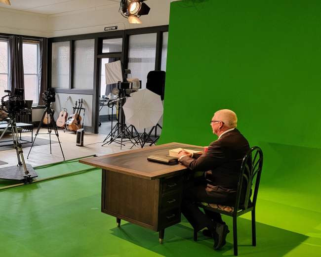 A man sits at a desk in front of a green screen in a brightly lit studio, surrounded by cameras and lighting equipment.