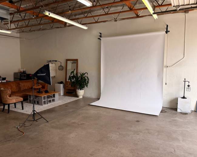 The image shows a spacious studio with a white backdrop, a brown couch, a mirror, a potted plant, and a light stand.