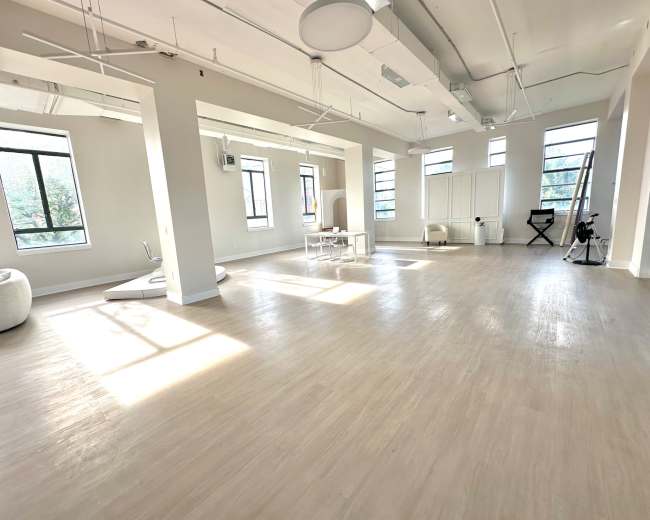 The image shows a spacious, well-lit room with hardwood floors, large windows, and minimal furniture, including a white table and chairs.