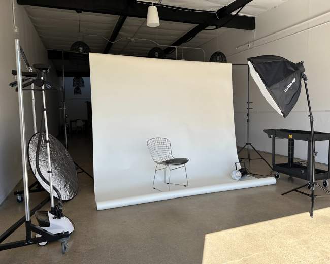 A photography studio features a white backdrop with a single chair in front, surrounded by lighting equipment and reflectors.