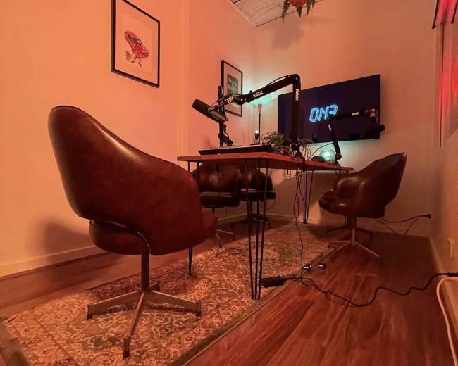 The image shows a small podcast studio featuring two brown chairs, a microphone setup on a table, and a screen displaying "ON3" on the wall.