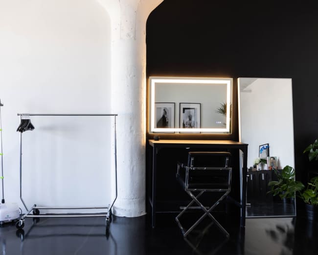 The image shows a modern salon interior featuring a clothing rack, a steam generator, a brightly lit mirror, and a chair against a dual-colored wall.