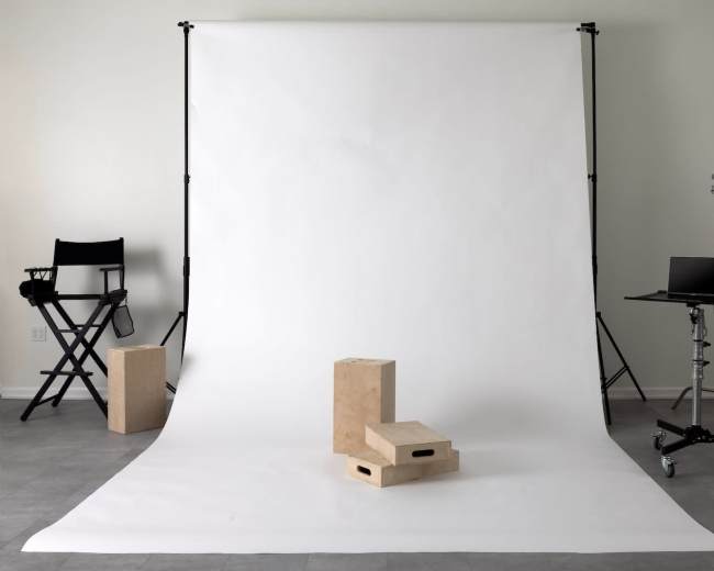 A photography studio with a white backdrop, two wooden boxes on the floor, a director's chair, and a mobile equipment cart.