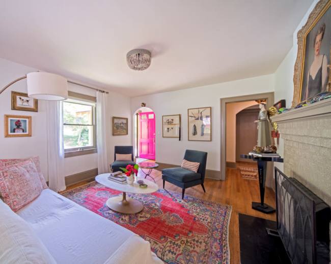 A bright and welcoming living room features a white sofa, two black chairs, a round coffee table, and colorful artwork on the walls, with a door painted bright pink.