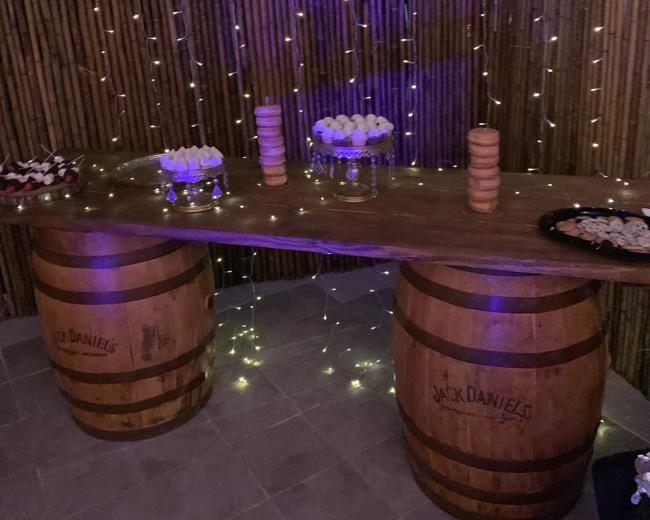A wooden table set on two barrels displays an assortment of desserts, with string lights hanging above in a dimly lit setting.
