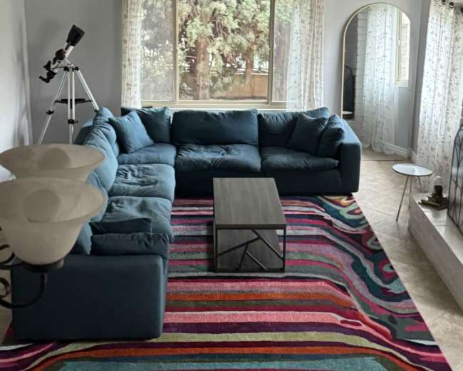The image shows a spacious living room featuring a large sectional sofa, a coffee table, and a colorful striped area rug, with tall windows allowing natural light to enter.