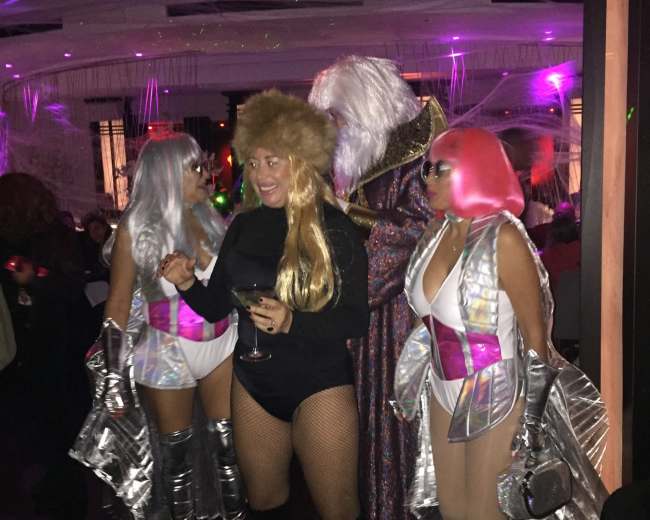 A group of four individuals, dressed in vibrant costumes with metallic accents, socializes in a colorful nightclub setting.