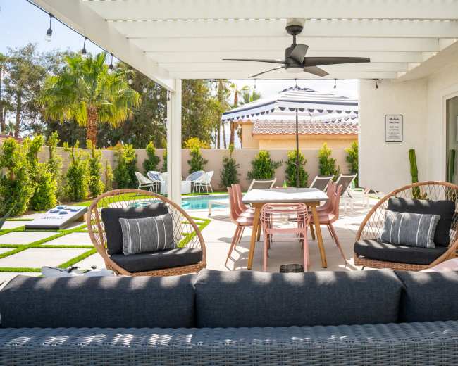 The image shows a patio area with seating, a dining table, and a swimming pool in a backyard surrounded by greenery.
