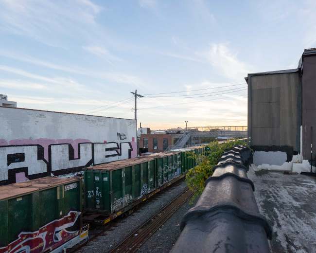 A view from a rooftop shows a line of green freight containers beside a railway, with a building and graffiti on the adjacent wall in the background.