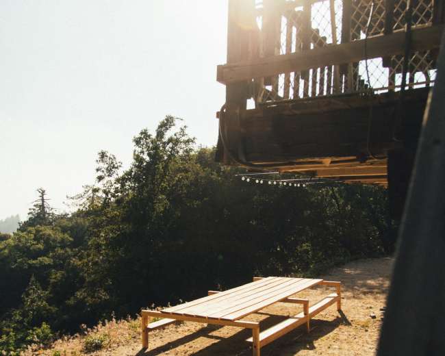 A wooden platform stands on a dirt area beneath a raised structure, with sunlight filtering through nearby trees.
