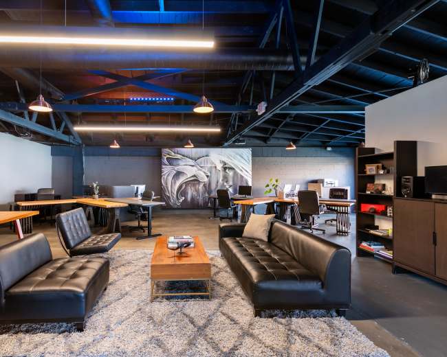 The image shows a modern office space featuring black leather sofas, a central wooden coffee table, and multiple workstations with computers and equipment in a well-lit environment.