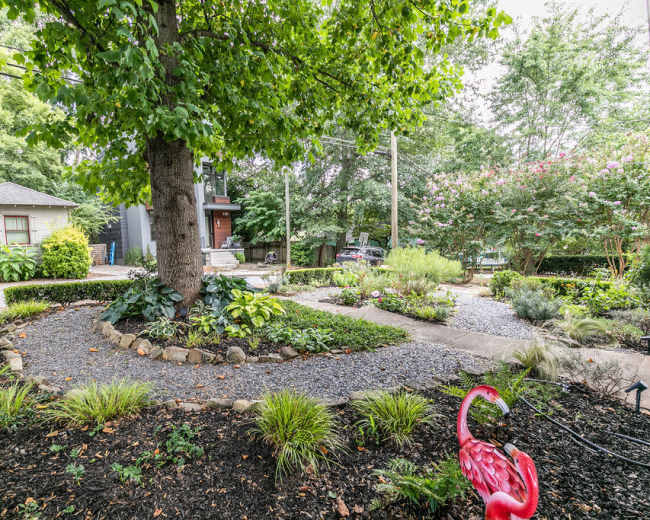 A landscaped garden features a large tree at its center, surrounded by gravel pathways and various plants, with two flamingo sculptures in the foreground.