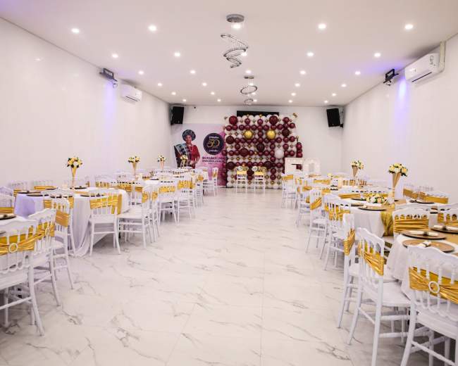 A spacious event hall is set up with white chairs and tables adorned with yellow tablecloths, featuring floral centerpieces, and a backdrop of balloons.