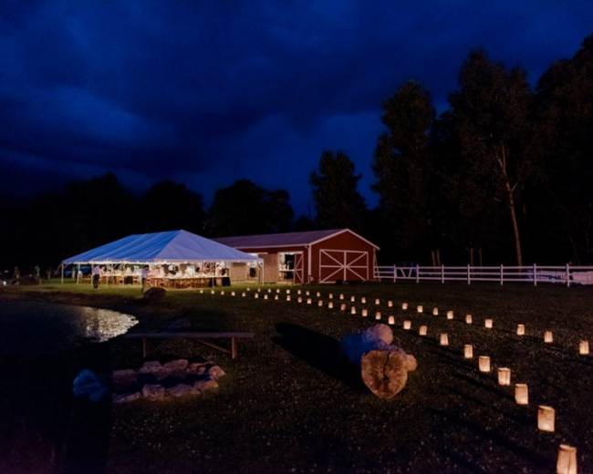 A large tent with lights is set up beside a pond and a red barn, illuminated by lanterns lining a pathway.