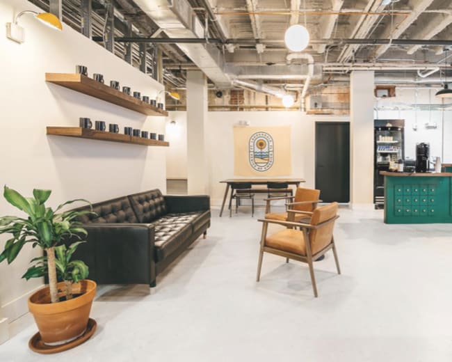 A modern office space featuring a black leather couch, wooden shelves with mugs, a green kitchen area, and a brown armchair.