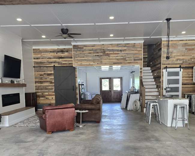 The interior of a modern open-concept home features a living area with a brown leather sofa, a kitchen with stainless steel appliances, and wooden wall accents.