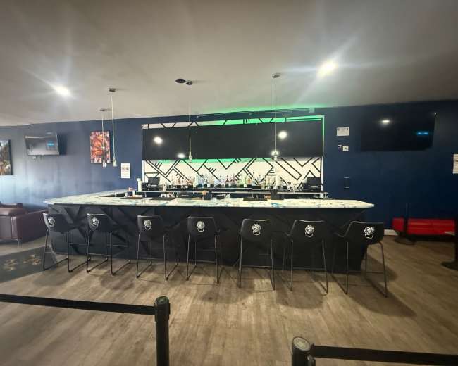 The image shows a modern bar area with a long marble-top counter, high black stools, and a brightly lit backdrop featuring various bottles and a geometric design.