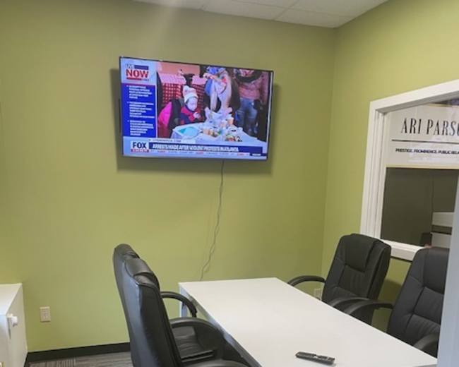 The image shows a small office meeting room with a white table, three black chairs, and a television mounted on the wall displaying a news program.