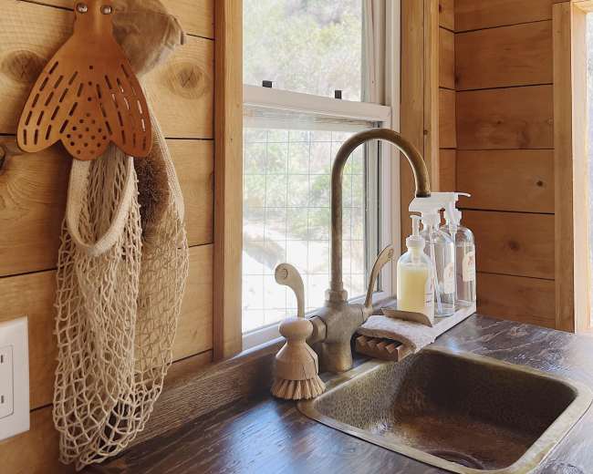 The image shows a kitchen sink with a bronze faucet, a dish scrubber, soap dispensers, and a netted bag hanging nearby, all set against wooden walls and a large window.