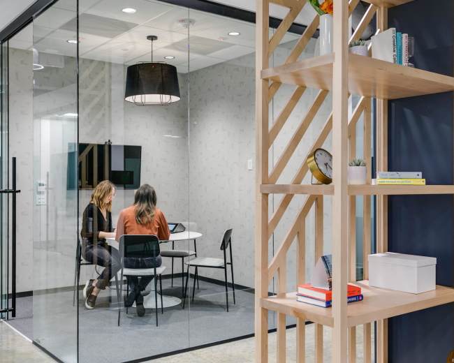 Two people are sitting at a round table inside a glass-walled conference room, while a wooden shelf with decorative items and books stands in the foreground.