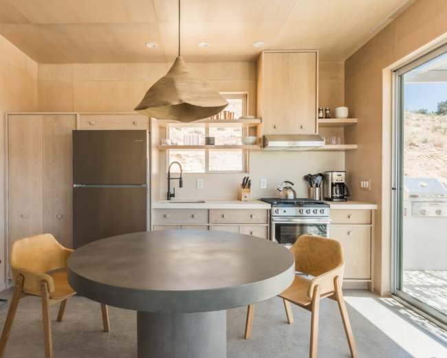 The image shows a modern kitchen with a round concrete table, two wooden chairs, a stainless steel refrigerator, a stove, and a sliding glass door leading outside.