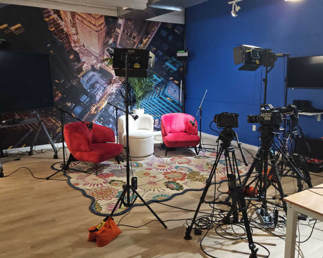 The image shows a studio setup with red chairs, multiple camera tripods, and lighting equipment, arranged in a space featuring a large urban-themed backdrop.