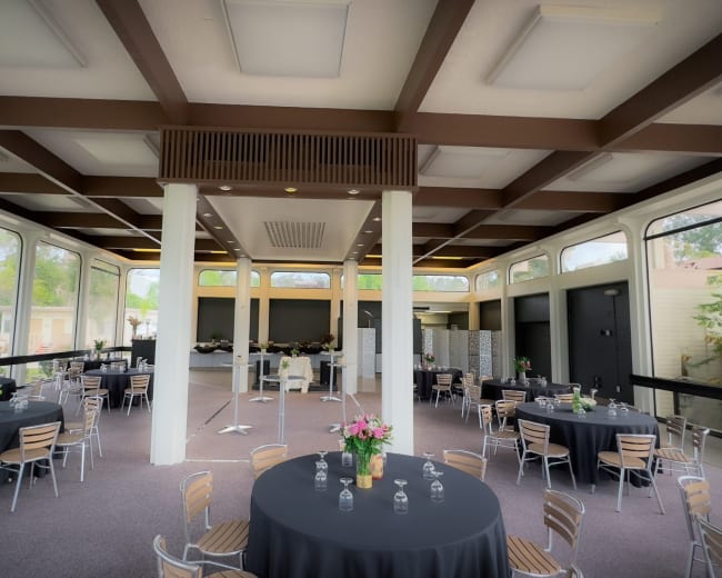 The image shows a spacious event hall with round tables dressed in black cloth, surrounded by large windows and a simple stage area at the back.