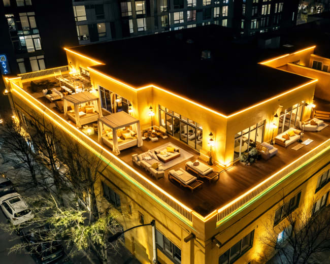 A rooftop terrace with outdoor seating is illuminated by warm string lights at night.