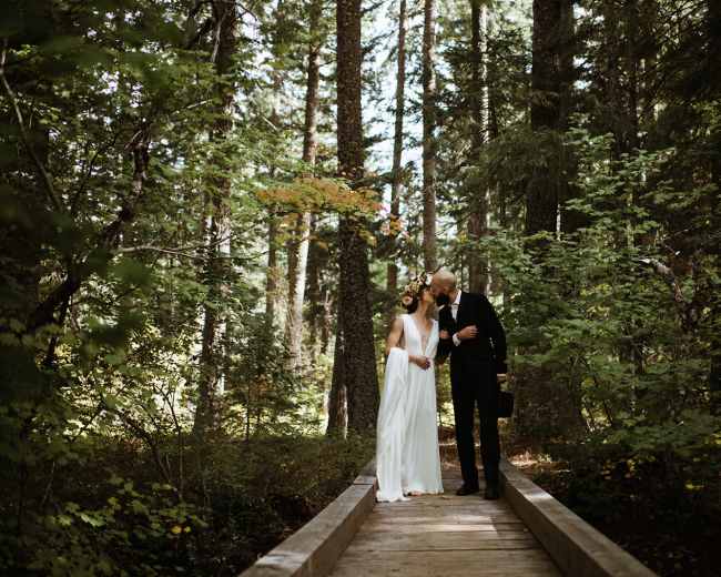 A couple in wedding attire stands close together on a wooden bridge, surrounded by trees and greenery.