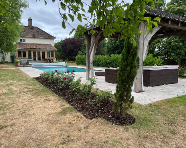 A well-maintained backyard features a swimming pool, a wooden gazebo, and neatly arranged flower beds alongside a two-story house.
