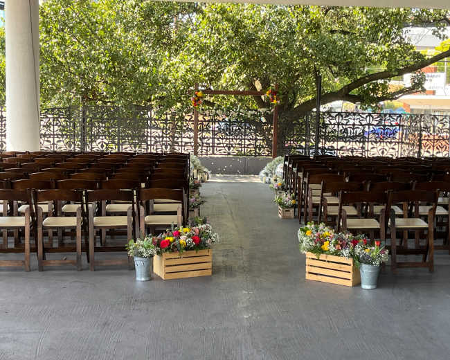 A wedding ceremony setup with rows of wooden chairs facing an arched structure decorated with flowers, framed by greenery.