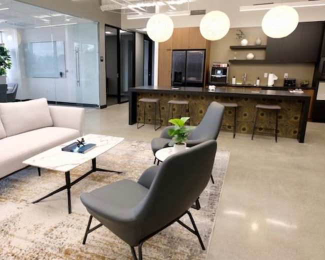A modern office lounge area with a sofa, two chairs, a coffee table, and a kitchenette in the background.
