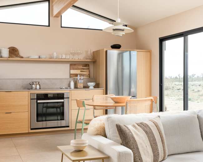 A modern kitchen and dining area features minimalist wood cabinetry, stainless steel appliances, and a round table with woven chairs overlooking a natural landscape through large glass doors.