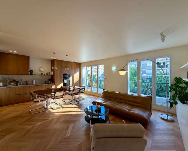 The image shows a modern living space featuring an open layout with a kitchen area, a dining table, a sofa, and large windows that provide natural light and a view of the outdoors.