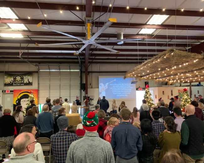 A large group of people stands in a warehouse-style space, facing a screen displaying text while festive decorations are visible around them.