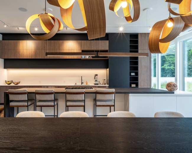 The image shows a modern kitchen with wooden pendant lights, a large dining table, bar stools at the counter, and sleek cabinetry.