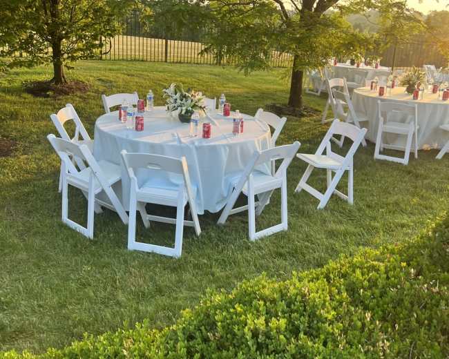 Several round tables with white tablecloths and chairs are set up on a grassy area, surrounded by trees and overlooking a field in the background.