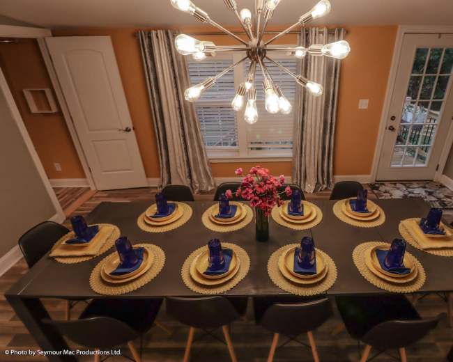A modern dining room features a long table set for twelve with gold chargers, blue glasses, and a floral centerpiece, under a large chandelier.