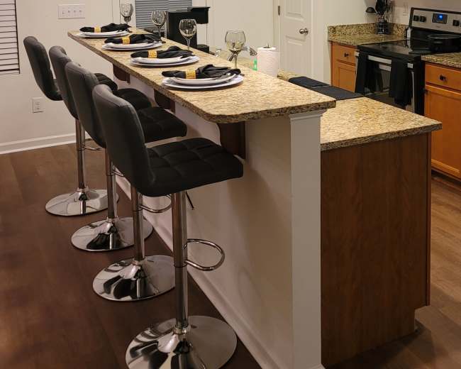 A modern kitchen features a bar area with four high stools, granite countertops, and pendant lights above, adjacent to the cooking area with wooden cabinets and stainless steel appliances.