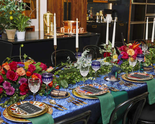 A long dining table is beautifully set with colorful floral arrangements, elegant glassware, and decorative plates, surrounded by candles and greenery.