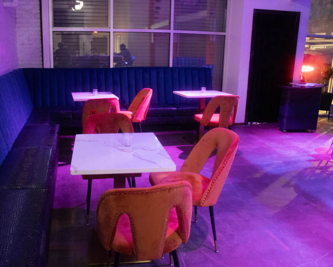 The image shows a modern lounge area featuring several upholstered chairs in orange and a blue couch, with white tables and colorful lighting.