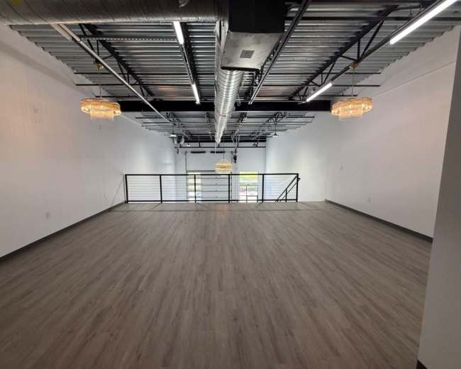 The image shows a spacious, empty room with light wood flooring, high ceilings, and modern lighting fixtures.